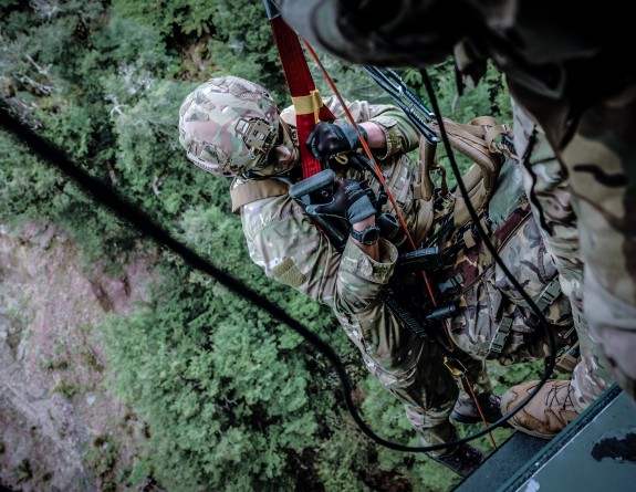 A New Zealand Army uniformed soldier winches down from a helicopter. The image is taken shown the helicopter loadmaster assisting the soldier down from the point of view looking down to the ground.
