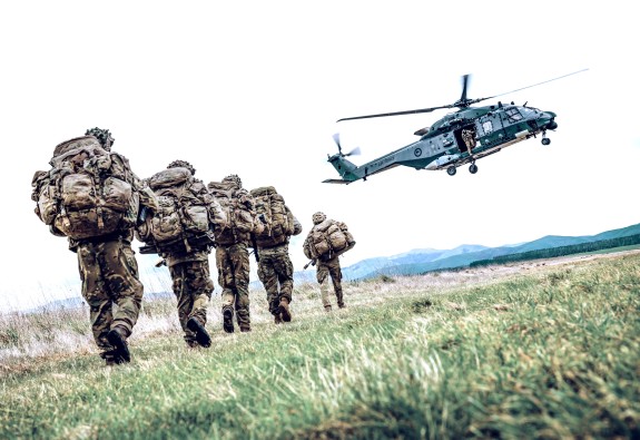 Uniformed soldiers walk toward an NH90 helicopter that's landing (hovering in the photo). The image has been shot from low down, on grass. 