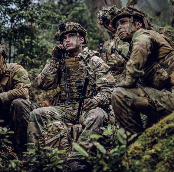 Uniformed soldiers in the bush. One is sitting looking up and holding a communications device, similar to a phone with cord. The other soldier next to them is also looking up.