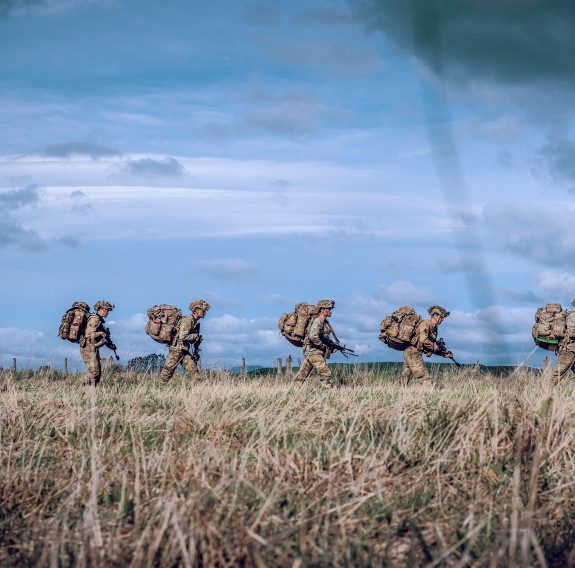 Uniformed New Zealand Army soldiers walk one after the other. The soldiers are wearing large packs, the sky is blue and it's a sunny day. The image is taken down in the grass - some shown in the foreground. 