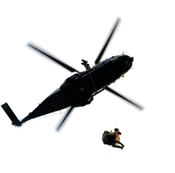An NH90 helicopter in the air while a soldier winches out of it. The photo is taken from right underneath, looking up. Cloudy skies. 