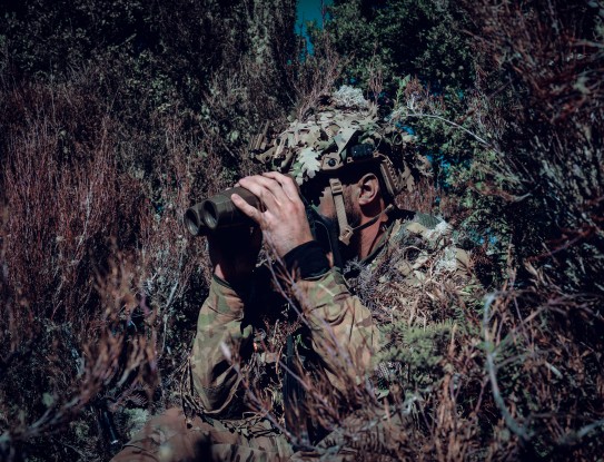 A New Zealand Army uniformed soldier blends in to their surroundings in the bush. They are looking to the left of the camera using binos. 