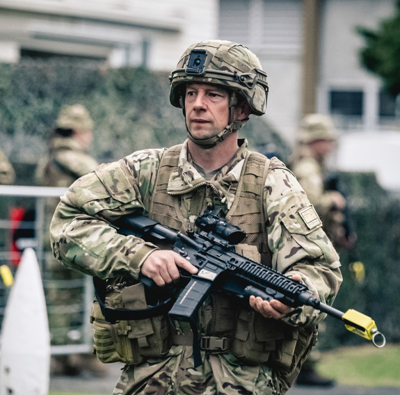 A soldier, holding a rifle looks to their right, in the background other soldiers are standing in front of a building.