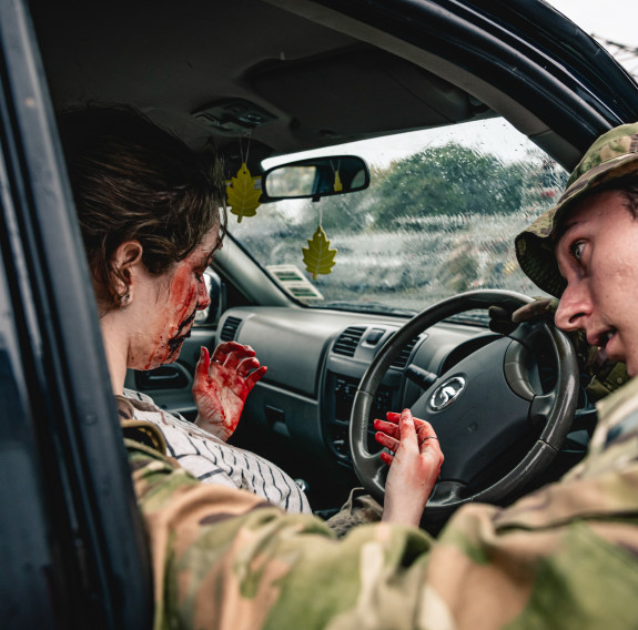 A driver sits in a vehicle covered in fake blood as a soldier attends to the mock medical event during an exercise.