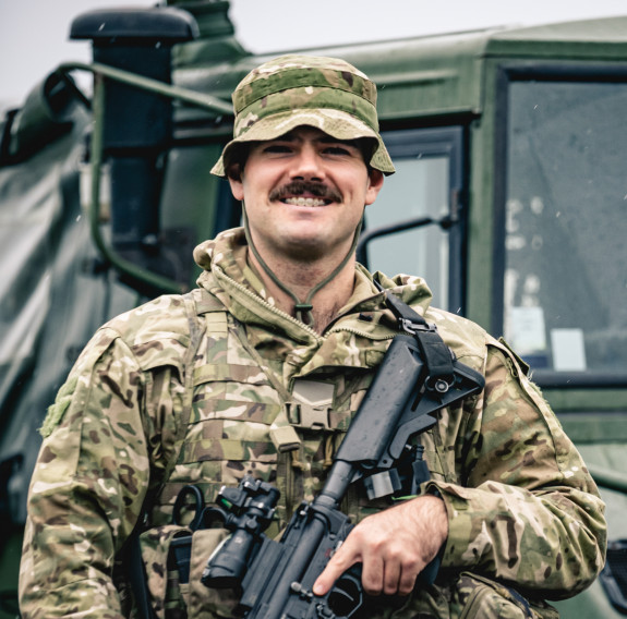 A soldier with a moustache holds a rifle while smiling at the camera. He is wearing camouflage Army uniform and standing in front of a dark green Army vehicle. 