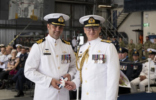 Two naval officers stand side by side in ceremonial uniform jointly clasping a bundled flag. Both officers stand in the foreground while an audience looks on at the rear, seated in an aircraft hangar.