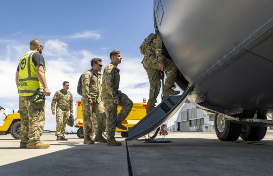 Military personnel use an aircraft's step to board the RNZAF Hercules. Under blue skies with scattered cloud, an aviator in high vis observes the boarding.