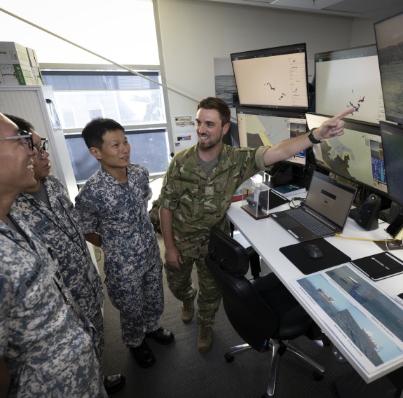 A man wearing forest camouflage uniform points to a wall of screens as three other personnel wearing grey and white camouflage uniform look on.