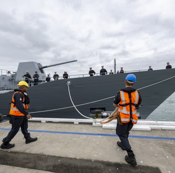 The sailors in orange high visability vests and hard hats untie a large grey ship from a dock. On the bow of the ship is a large gun, a New Zealand flag and sailors.