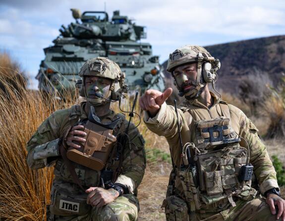 NZ Army JTAC personnel pointing toward the camera and look like they are talking, they have comms on. In the background is an NZLAV. They are in a grassy, tussock area. 