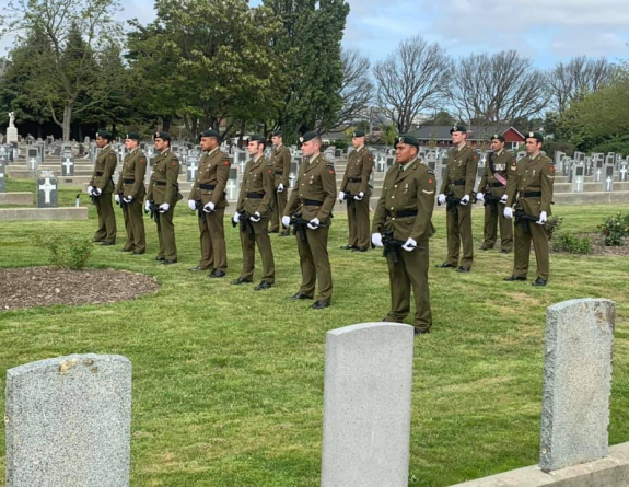 Soldiers stand in formation with their weapons in hand on a grassed area of a cemetery. 