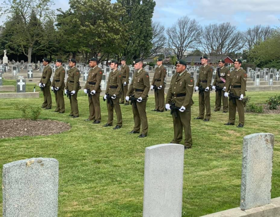 Soldiers stand in formation with their weapons in hand on a grassed area of a cemetery.