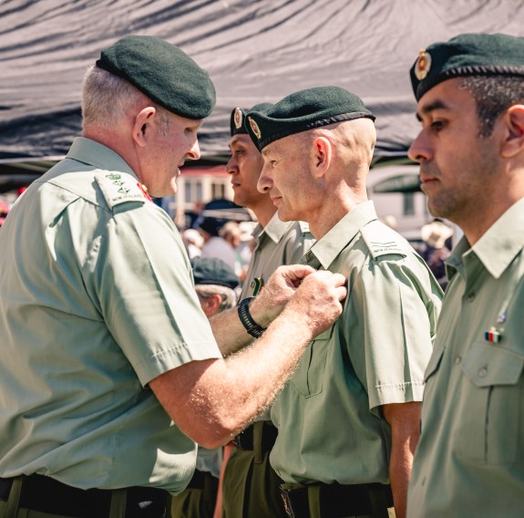 A parade of soldiers stand at attention, whilst one officer pins a badge to one of the soldiers adorned with green shirts and berets. Marquees can be pictured in the background filled with guests of the event.