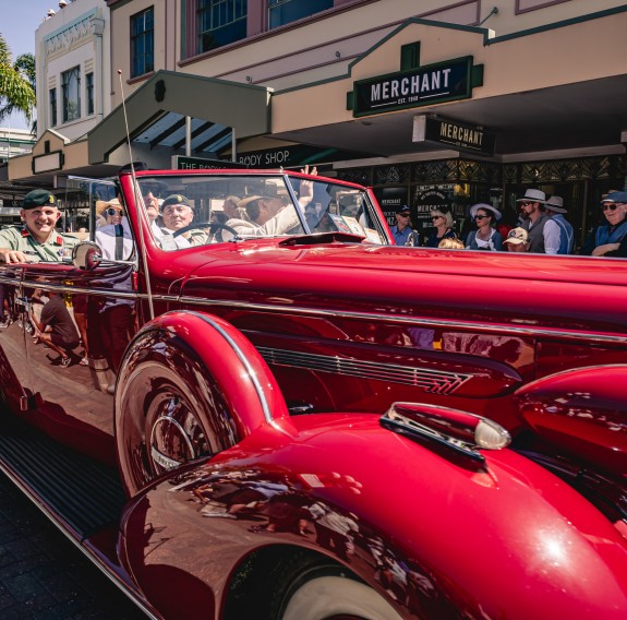 A large red vehicle drives down a street flanked with members of the public dressed in art deco era clothing, the vehicle holds army staff smiling and waving at the crowds.