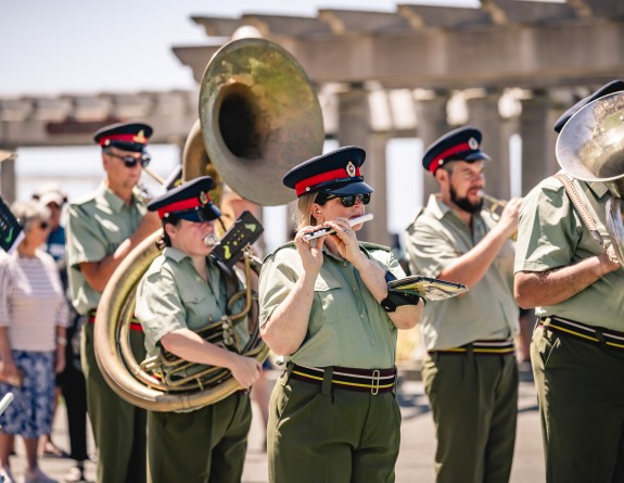 An army band assembles at the esplanade dressed in green shirts and trousers playing various brass instruments.