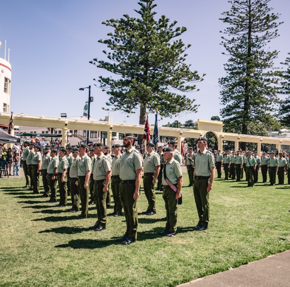 A parade of army soldiers takes part on a grassed area in front of a long boulevard and art deco architecutre.