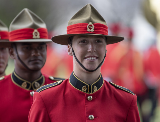 A woman smiles while standing in formation with other graduating Officers. She is wearing a brown and red lemon squeezer (type of hat) with a neck strap and a red Army jacket with black and gold accents.