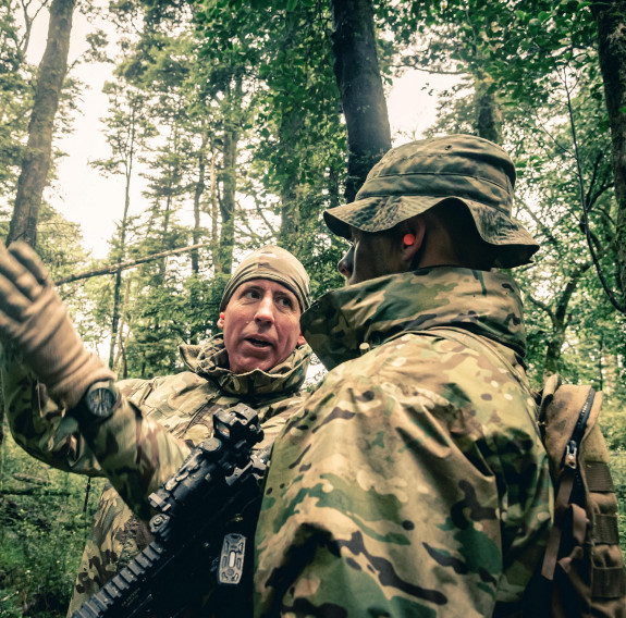 A solider explaining something to another soldier amongst tall trees.