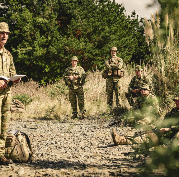 Soldiers sit and stand around a gravel area as a soldier is talking to the group.