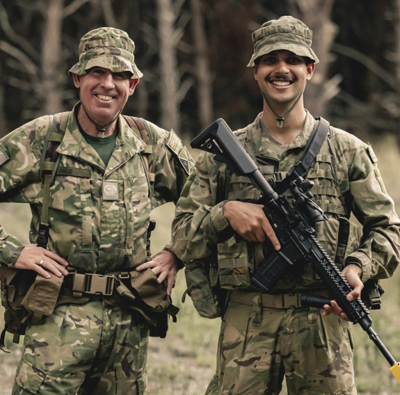 Two soldiers smile. The man on the left has his hands on his hips, while the man on the right holds a rifle.