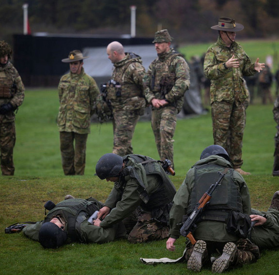 Soldiers stand in the background and soldier in the foreground administer first aid.