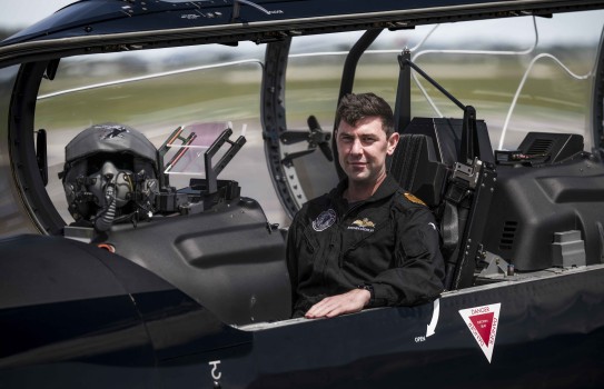 A male wearing a black flying suit sits inside the cockpit of a small aircraft with the hood open. His hand rests on the side of the aircraft whilst looking at the camera.