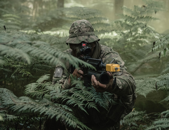 A soldier crouches in the bush, carrying a weapon. There are native ferns around them. 
