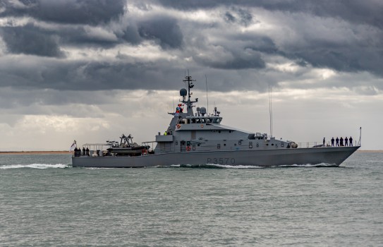 A grey military vessel cruises on the ocean whilst a smaller white and orange boat follows behind in its wake.