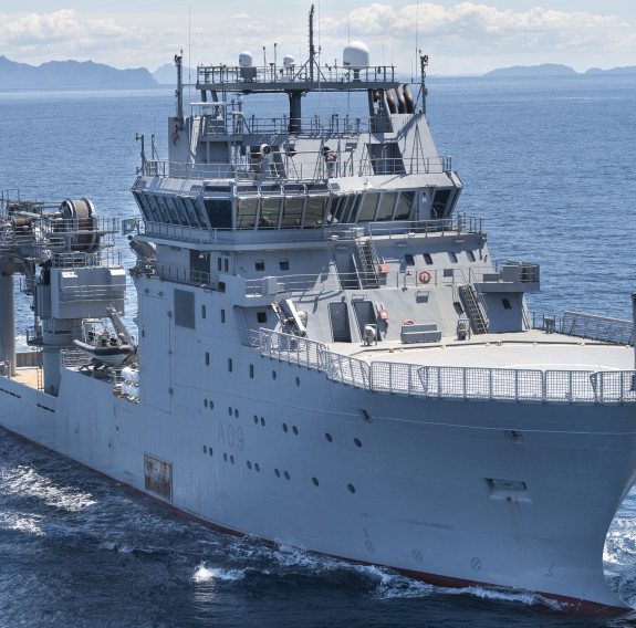 Stock photo of HMNZS Manawanui gliding through the ocean on a sunny day with land and hills in the distance. The ship is grey and rather bulky with a large crane on the back.