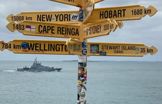 A yellow signpost located in Bluff points to multiple locations, from Stewart Island to Cape Reinga, to the Equator, New York and Hobart. The sea is visible in the background with a grey military vessel cruising near the horizon.