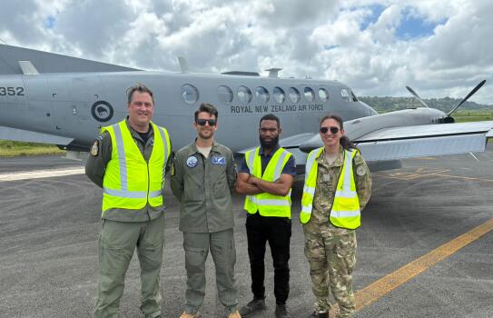 Four people, three in high vis, stand in front of a small grey aircraft under clouds on the flight line.