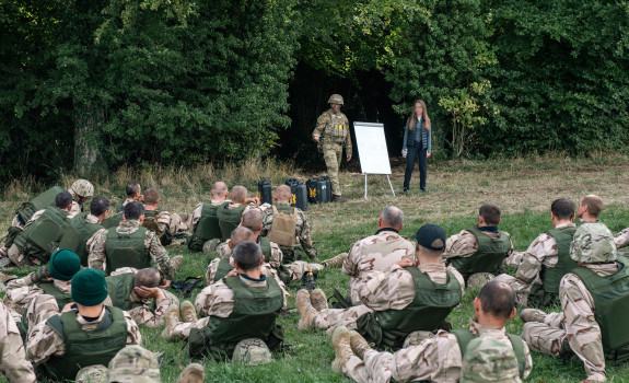 A soldier and a woman stand next to a white board while talking to a group of soldiers sitting at the edge of a field, by the trees.