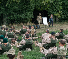 A soldier and a woman stand next to a white board while talking to a group of soldiers sitting at the edge of a field, by the trees.