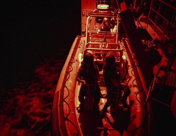 A naval boat is alongside a large naval ship at night time with several people wearing helmets and uniform inside.