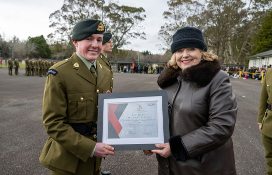 Angus, in Army uniform (left) accepts a certificate from Minister of Defence, Judith Collins (right), who is wearing a brown leather jacket with a fur collar and a black hat on the parade ground during prize giving.