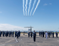 Royal New Zealand Air Force personnel standing in a formation on the flight line on a nice blue sky day. Five T-6C Texan II aircraft fly overhead and a King Air 350 aircraft sits in the background.