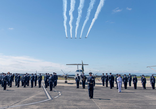 Royal New Zealand Air Force personnel standing in a formation on the flight line on a nice blue sky day. Five T-6C Texan II aircraft fly overhead and a King Air 350 aircraft sits in the background.