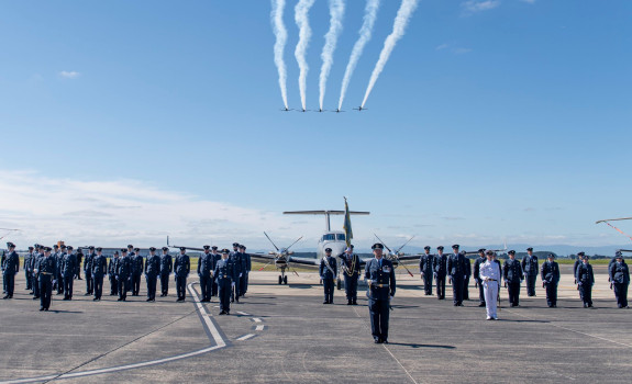 Royal New Zealand Air Force personnel standing in a formation on the flight line on a nice blue sky day. Five T-6C Texan II aircraft fly overhead and a King Air 350 aircraft sits in the background.