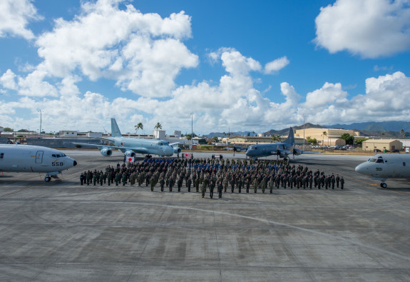 A group of personnel standing in front of various aircraft. The sky is blue with some clouds.