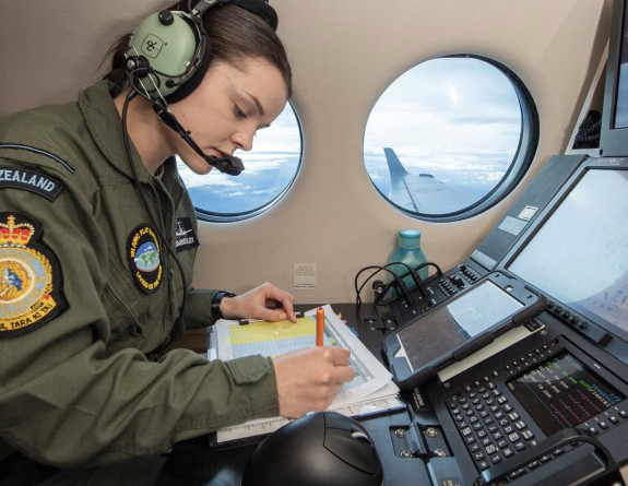 A Royal New Zealand Air Force Air Warfare Officer wearing a headset documents at a station in the King Air 350 aircraft during flight.