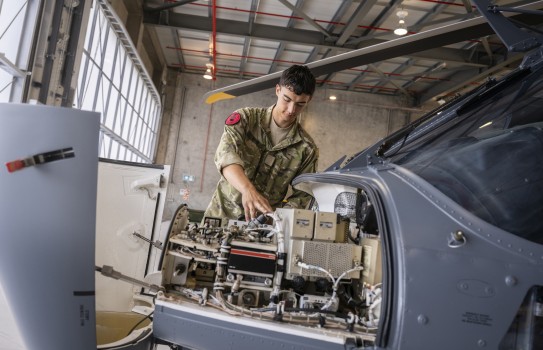 An aviator works on the engine on a grey helicopter in an aircraft hangar.
