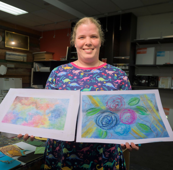 Woman smiles as she holds two colourful works of art made at the workshop.