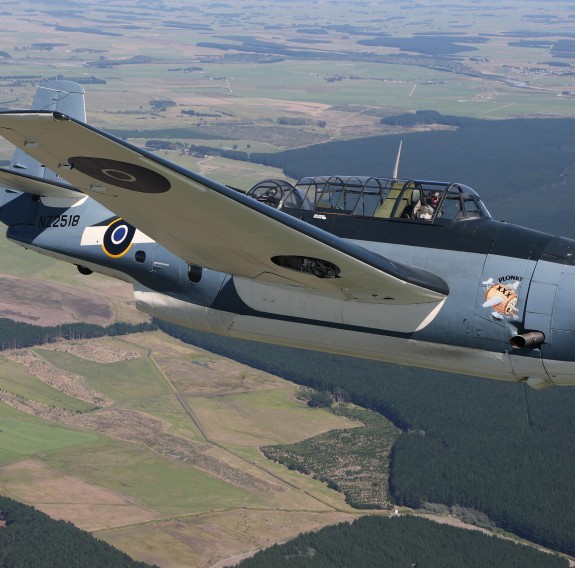 A historic small aircraft flies through the sky with farmland as far as the eye can see below.