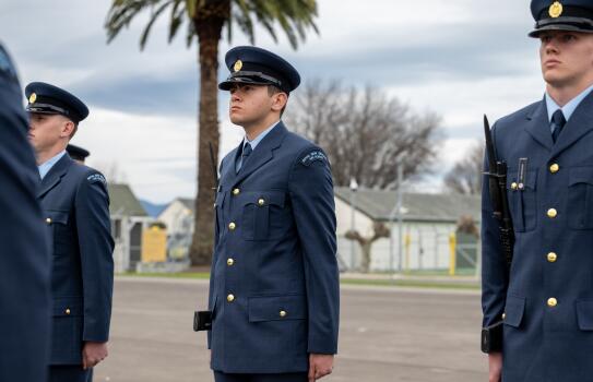 An aviator stands to attention on the a parade ground alongside other aviators. In the background are military buildings and a large palm tree.