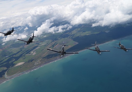 A formation of five aircraft fly high in the blue sky above the sea and land below.