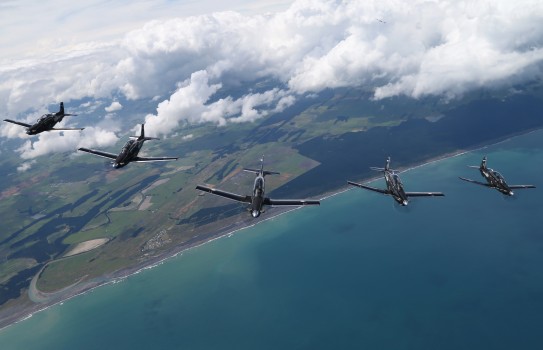 A formation of five aircraft fly high in the blue sky above the sea and land below.