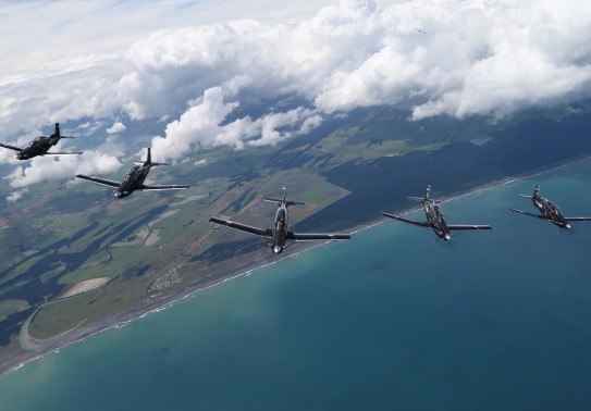A formation of five aircraft fly high in the blue sky above the sea and land below.