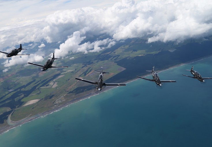 A formation of five aircraft fly high in the blue sky above the sea and land below.