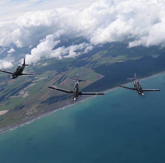 Five black aircraft fly in formation above the clouds, sea and land.