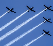 A formation flight of five aircraft in the shape of an arrowhead soars in the blue sky with white trails behind.